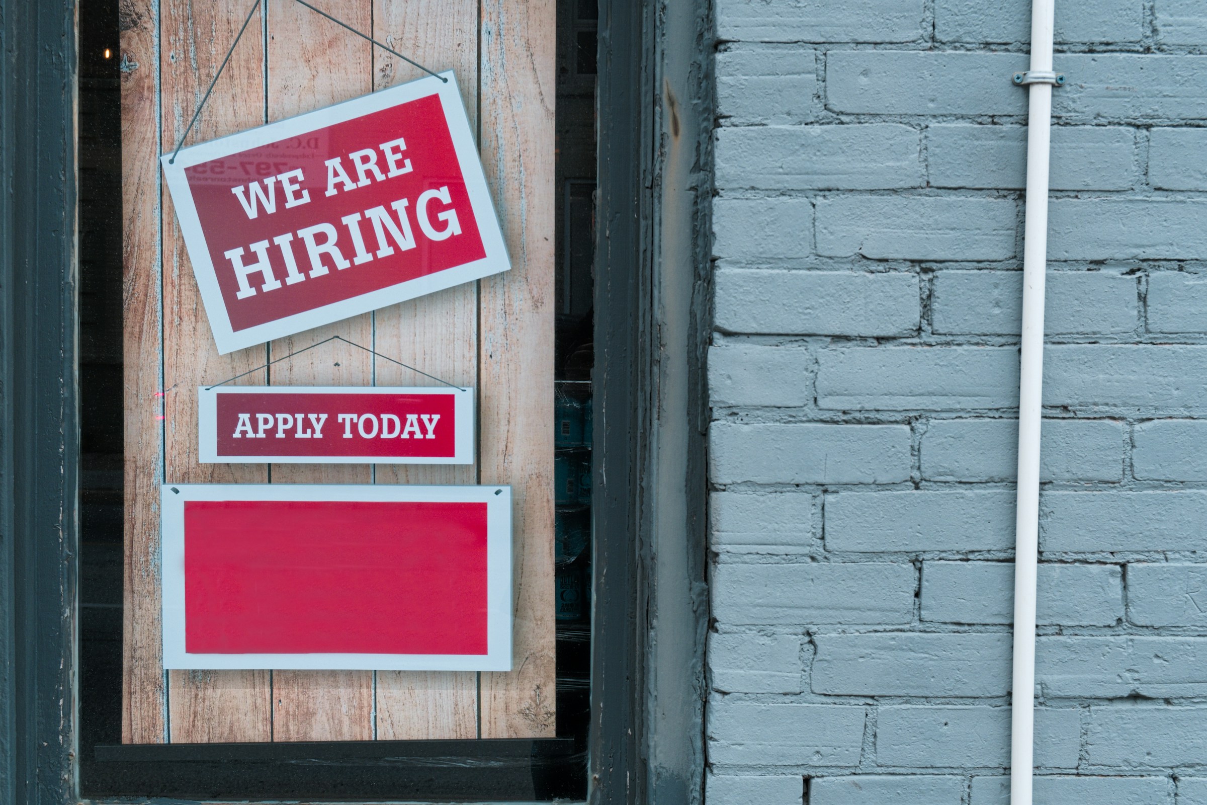 Schaufenster mit drei roten und weißen Schildern: Oben steht 'WE ARE HIRING', darunter 'APPLY TODAY', das unterste Schild ist leer.