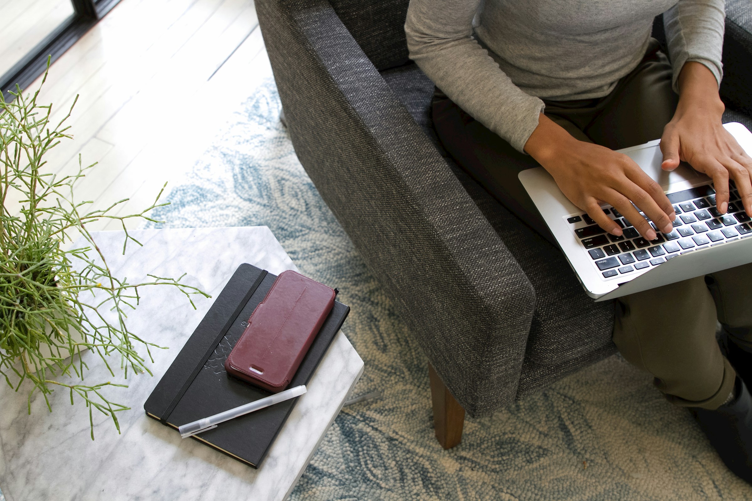 Person sitzt in einem grauen Sessel und tippt auf einem Laptop. Daneben steht ein kleiner Marmortisch mit einem Notizbuch, einem Smartphone in roter Hülle und einem Stift. Im Hintergrund ist ein Teppich mit blau-weißem Muster und eine grüne Pflanze sichtbar.