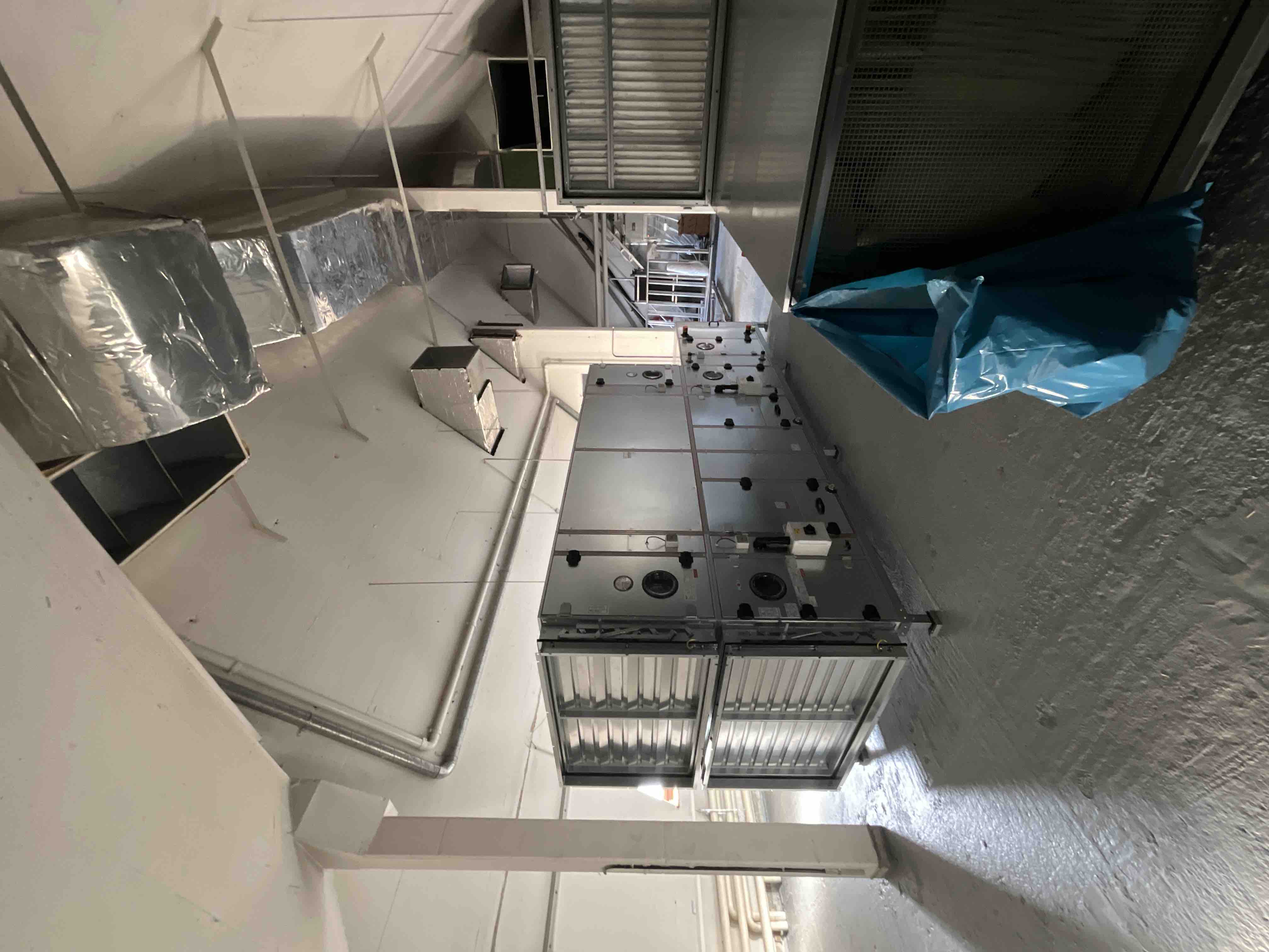 Installierte Lüftungsanlage mit großen Metallgehäusen und Luftfiltern in einem Technikraum. Sichtbar sind Rohrleitungen und Kanäle für Heizungs-, Lüftungs- und Klimatechnik (HLK), ideal für industrielle Gebäudetechnik und professionelle Luftaufbereitungssysteme.