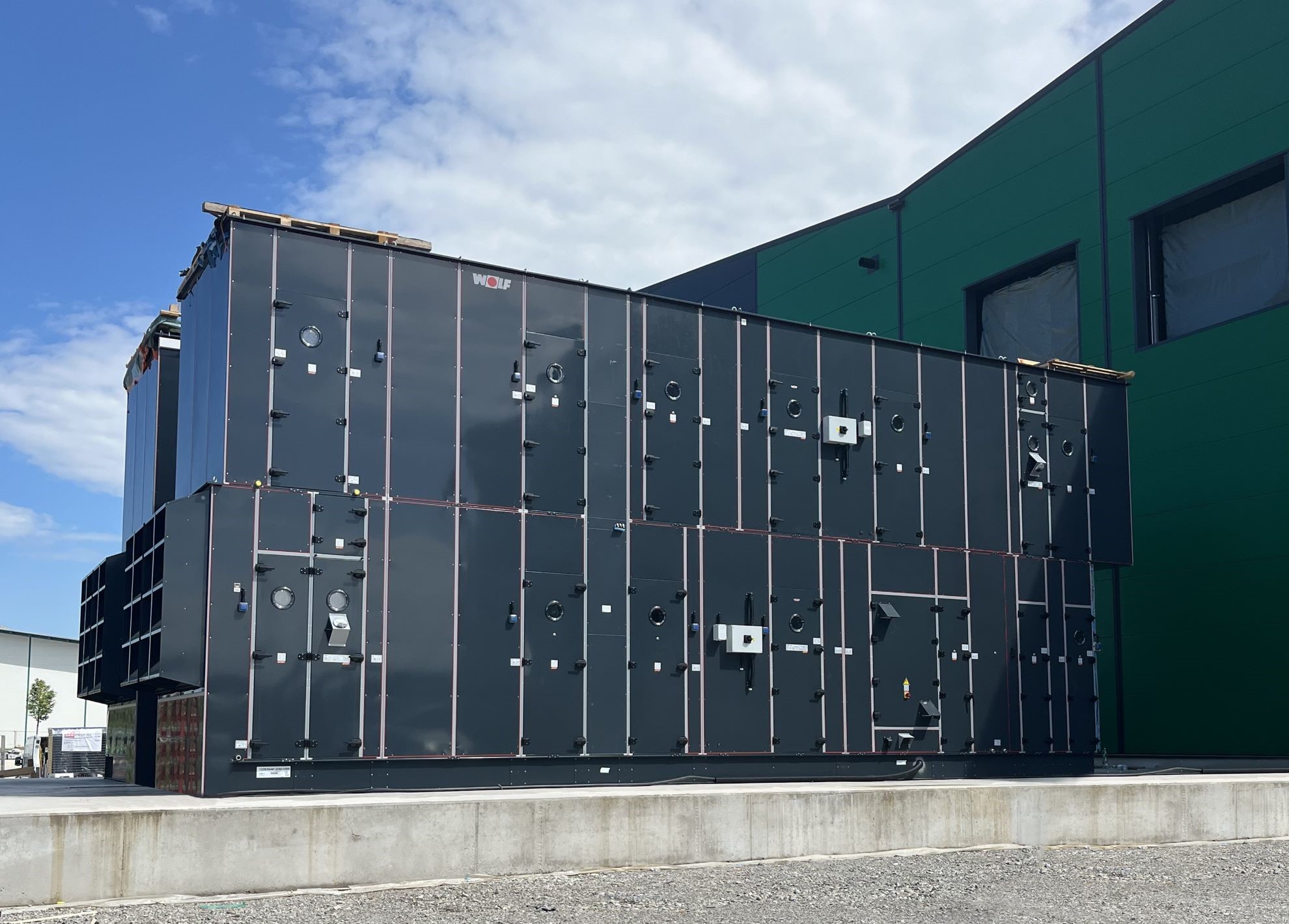 Große Lüftungsanlage in schwarzer Ausführung auf einem Betonfundament vor einem Industriegebäude. Die Anlage ist für Heizungs-, Lüftungs- und Klimatechnik (HLK) konzipiert und wird im Bereich Gebäudetechnik und industrielle Lüftung eingesetzt.