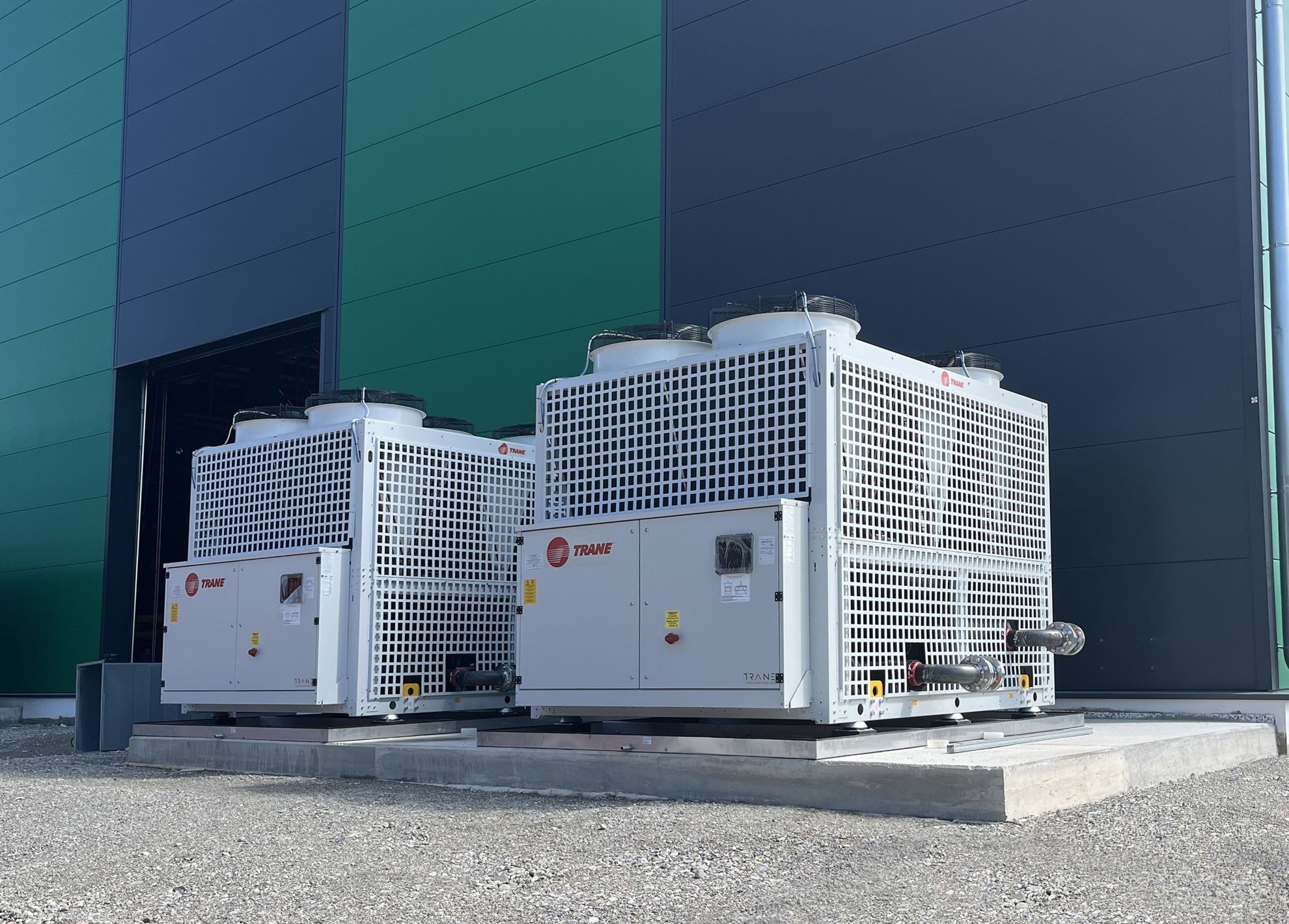 Zwei große Wärmepumpen auf Betonfundamenten vor einem Industriegebäude. Die Geräte sind für Heizungs-, Lüftungs- und Klimatechnik (HLK) ausgelegt und dienen der energieeffizienten Gebäudetechnik sowie industriellen Kühlung und Heizung.